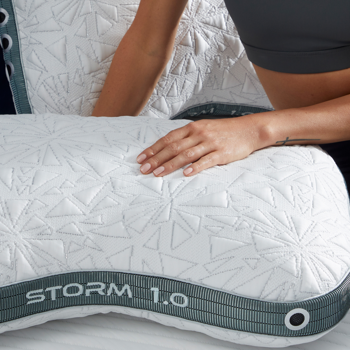 A close-up of a white quilted Storm 1.0 pillow with a gray branded trim, shown with a person's hand resting on it and part of their torso visible.