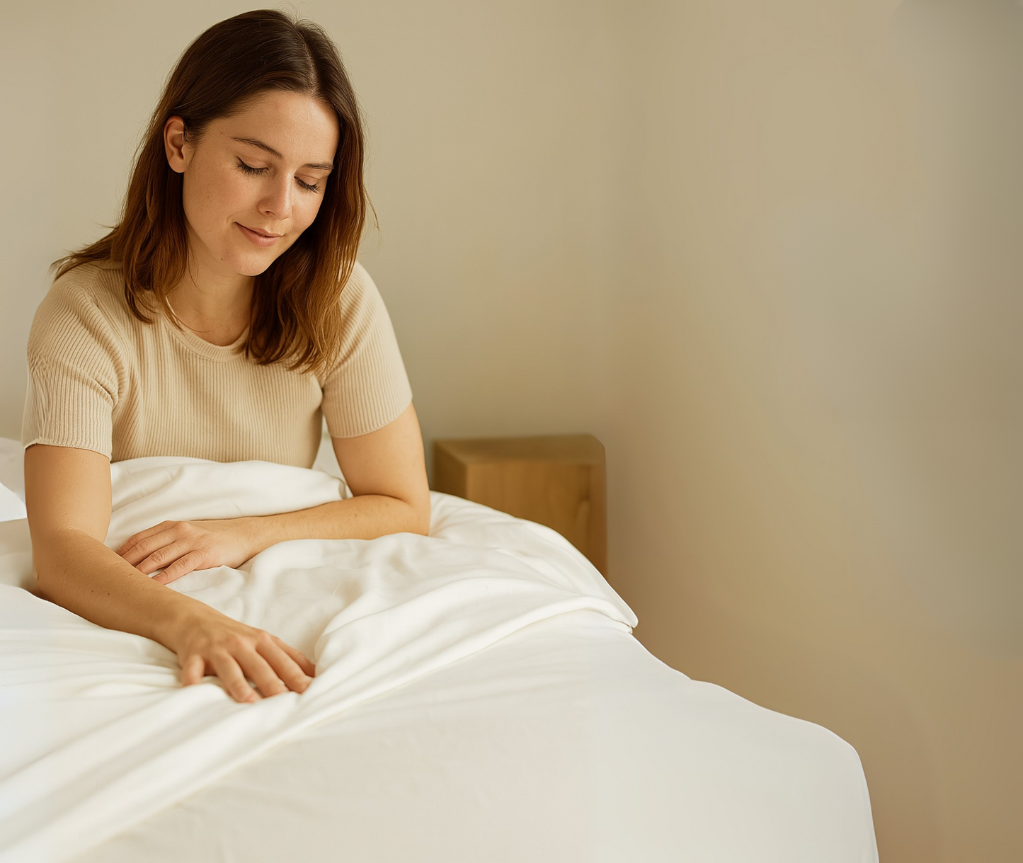 Woman sitting on a bed with white bedding in a softly lit room.